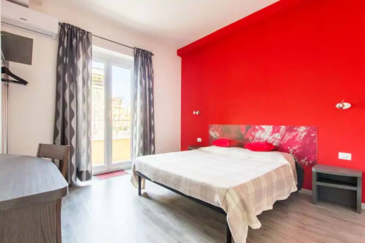Un dormitorio con cama, una mesita, una silla, una mesita de noche y una pared de acento roja.