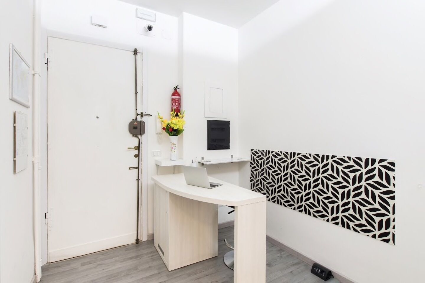A room with a white door, a white desk with a laptop, a fire extinguisher, and a patterned wall panel.