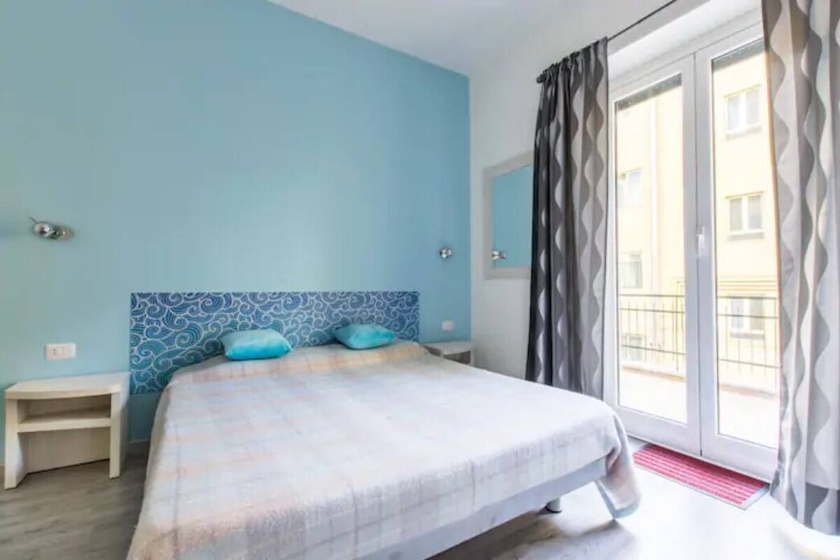 Un dormitorio con cama, mesita de noche, ventana con cortinas y vista a edificios.