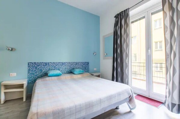 A bedroom with a bed, a nightstand, a window with curtains, and a view of buildings outside. - B&B di Paolo S. (Rome)