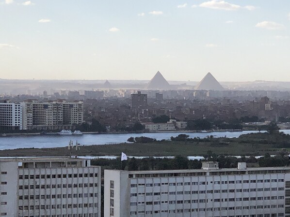 A cityscape with two prominent pyramids in the background, a river in the foreground, and several multi-story buildings. - Three Bedrooms Apartment Nile view Maadi (Maadi)