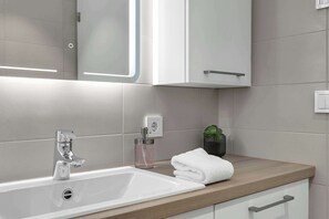 Une salle de bain moderne avec un lavabo blanc, un robinet chromé et un plan de toilette en bois. On y trouve une serviette pliée sur le plan de toilette et une petite plante en pot.