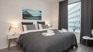A modern hotel room with a large bed, a nightstand with a lamp, a chair, and a window with curtains. - SIF Apartments Central Reykjavik - 1 Bedroom (Reykjavík)