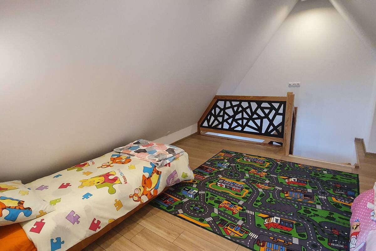 A small, cozy room with a single bed featuring a colorful cartoon character print, a play mat with a road and vehicle design, and a wooden ladder against the wall.