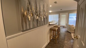 A hallway with a gray abstract wall art, a white door, and a kitchen area with a counter and stools.