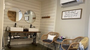 Un baño con lavamanos, espejo y aire acondicionado instalado en la pared. Hay sillas de mimbre y una mesita con botellas.