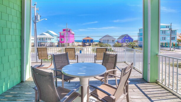 Ein überdachter Außenbereich mit Tisch und Stühlen bietet Blick auf bunte Strandhäuser und den Ozean.