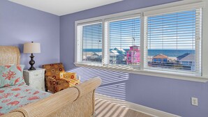 Ein Zimmer mit großem Fenster, einem Bett mit floralem Bettbezug, einer Lampe und einem Korbstuhl.