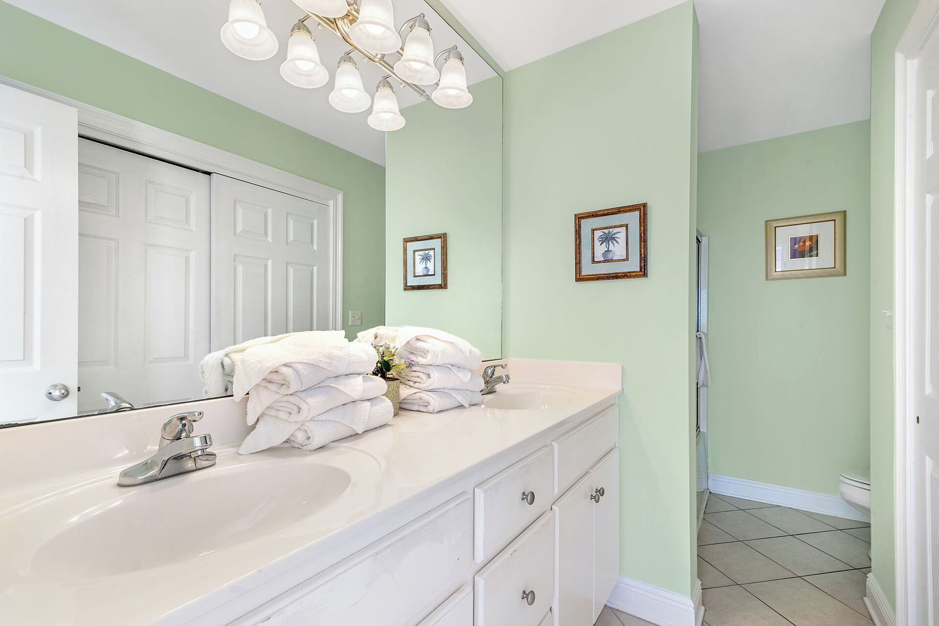 A bathroom with a white vanity, a large mirror, and a light green wall. There are framed pictures on the wall and a stack of white towels on the vanity.
