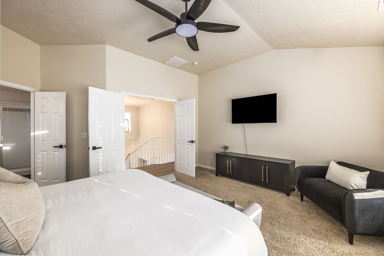 A bedroom with a bed, a TV mounted on the wall, a ceiling fan, and a staircase leading to another room.