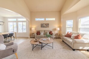 A spacious living room with a high ceiling, a beige sofa, two round coffee tables, and a dining area with a wooden table and chairs.