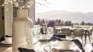 Une table à manger dressée avec de l’assiette blanche, des bols et des verres à vin, un vase blanc orné de branches, et une vue sur des montagnes en arrière-plan.