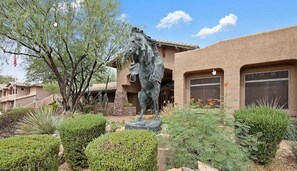 Un edificio con una gran estatua de un caballo al frente, rodeado de jardines cuidados y un cielo azul despejado.