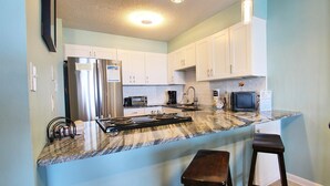 A kitchen with white cabinets, a stainless steel refrigerator, a microwave, a stove, and a small television. There are two stools and a countertop with a granite-like backsplash. - Compass Point 205 (Gulf Shores)