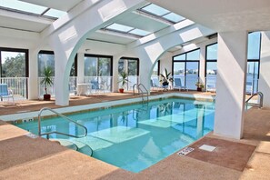 Une piscine intérieure à l’eau d’un bleu cristallin, entourée de colonnes blanches et de grandes fenêtres.