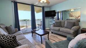 A living room with a sofa, a coffee table, and a television. There is a balcony with a view of the water. - Compass Point 205 (Gulf Shores)