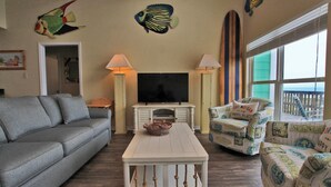 A living room with a gray sofa, a white coffee table, and two armchairs with beach-themed cushions. A flat-screen TV is on a white cabinet, and there are two lamps on either side. - Cabana Beach House (Gulf Shores)