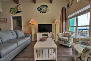 A living room with a gray sofa, a white coffee table, and two armchairs with beach-themed cushions. A flat-screen TV is on a white cabinet, and there are two lamps on either side.