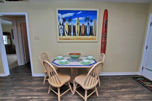 A dining area with a table set for four, surrounded by wooden chairs. A colorful painting of surfboards is on the wall above the table. - Cabana Beach House (Gulf Shores)