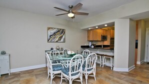 A dining area with a glass table and white wicker chairs, a ceiling fan, and a kitchen with wooden cabinets and appliances. - Broadmoor 304 (Orange Beach)