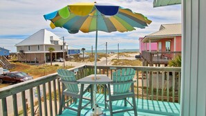 A wooden deck with a table and chairs, a colorful umbrella, and a view of the beach and houses. - Dream Catcher (Gulf Shores)