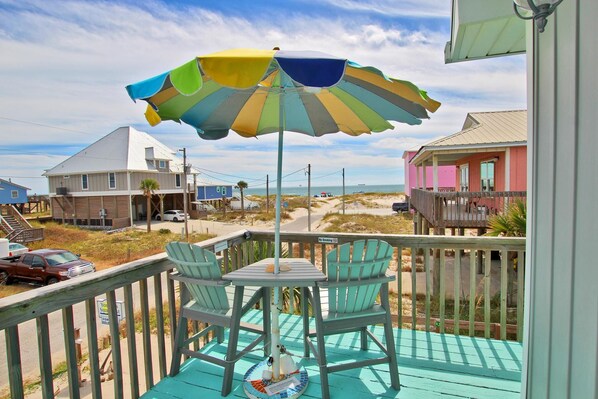 A wooden deck with a table and chairs, a colorful umbrella, and a view of the beach and houses.