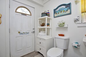 A small, clean bathroom with a white toilet, a white sink, and a white vanity with drawers. There is a shelf above the toilet with a decorative item and a basket. A sign on the door reads "Keep Door Closed to Maintain Set Temperature."