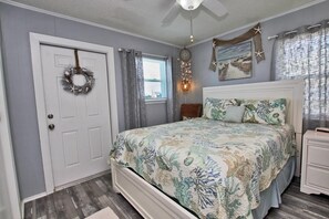 A bedroom with a bed, a nightstand, a dresser, a door with a wreath, a window with curtains, and a wall with a dreamcatcher and starfish decoration.