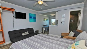 A bedroom with a large bed, a ceiling fan, a TV mounted on the wall, and a view into the kitchen area. - Beachview 203 (Gulf Shores)