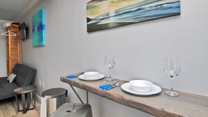 A modern room with a wooden dining table set for two, a grey sofa, a small round table, and a painting of a seascape on the wall. - Beachview 203 (Gulf Shores)