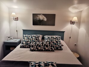 Una cama bien hecha con almohadas estampadas, dos lámparas fijadas a la pared y un cuadro enmarcado sobre la cama.