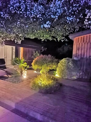 Un espacio al aire libre iluminado de noche, con terraza de madera, plantas variadas y una pequeña fuente.