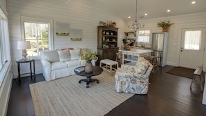 A spacious living room with a white sofa, a floral armchair, and a coffee table. There is a dining area with a wooden table and chairs, and a kitchen area with a refrigerator and a sink.
