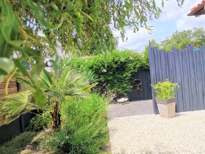 A garden area with a gravel path, a wooden fence, and various plants including a palm tree and flowering shrubs. - STUDIO "house" with TERRACE at the foot of the cycle paths (Audenge)