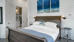 A bedroom with a wooden bed, white bedding, a nightstand, a wall-mounted lamp, and a large window with a view of the ocean. - Stay@‘Summer Breeze,’ a 2-Story, Cinnamon Shore Townhome w/ Great Amenities (Port Aransas)