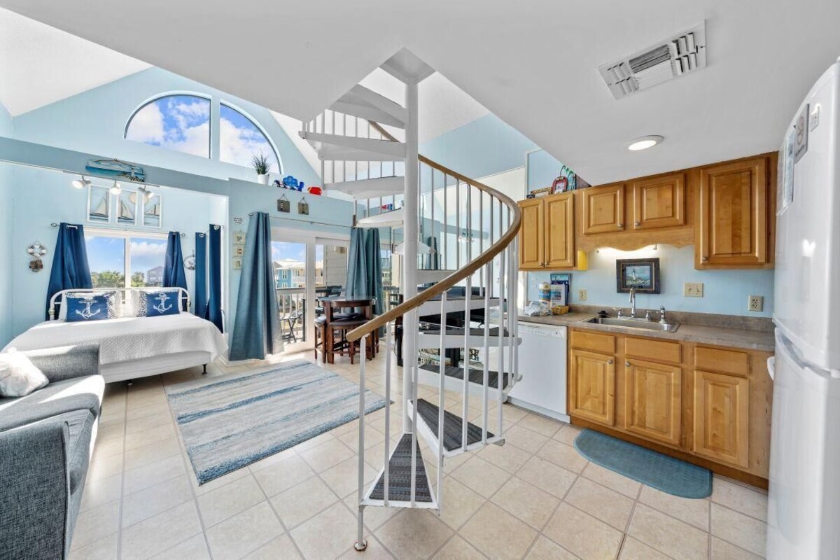 A spacious living area with a large bed, a staircase leading to an upper level, a kitchen with wooden cabinets, and a view of the sky through a skylight.