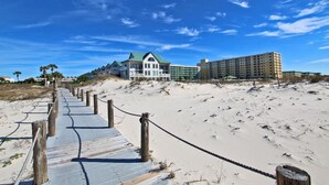 A beachside hotel with a wooden boardwalk leading to the beach, featuring a large building with a green roof and a smaller building with a white facade.