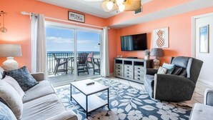 A living room with a sofa, armchair, coffee table, and a flat-screen TV. There's a balcony with a table and chairs, and a ceiling fan. - Legacy 102 (Gulf Shores)
