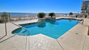 A rectangular pool with a metal ladder, surrounded by a concrete deck. There are lounge chairs and a palm tree on the deck. A fence separates the pool from the beach. - Legacy 102 (Gulf Shores)