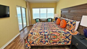 A bedroom with a large bed, a ceiling fan, a TV, and a view of the ocean through the window.