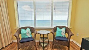 Two wicker chairs with cushions, a small round table, and a view of the ocean through large windows.