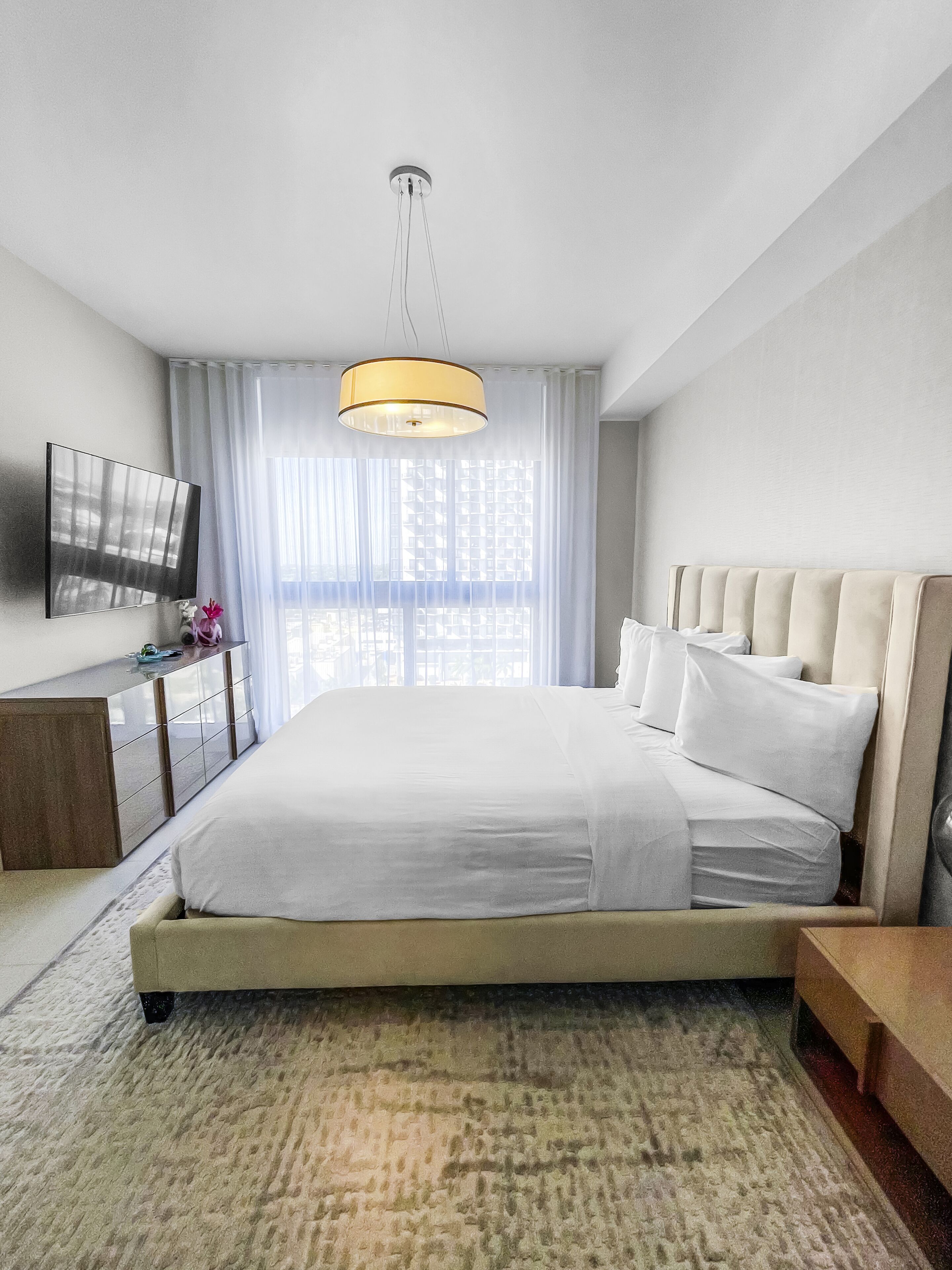Une chambre d’hôtel moderne dotée d’un grand lit, d’un téléviseur à écran plat fixé au mur, d’une table de chevet et d’une grande fenêtre avec des rideaux légers.