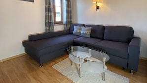 Un salon moderne avec un canapé d’angle aux couleurs sombres, une table basse en verre et une lampe fixée au mur.
