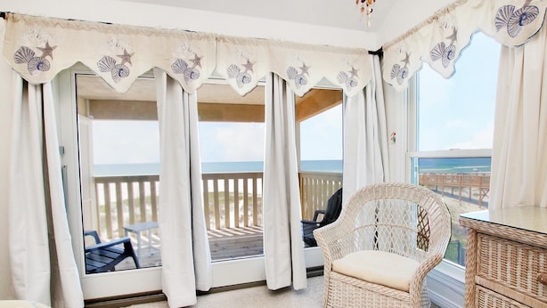 Un balcon avec un fauteuil en osier, des rideaux blancs ornés de motifs d’étoiles de mer et de coquillages, et une vue sur l’océan.