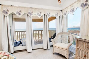 Ein Balkon mit einem Korbstuhl, weißen Vorhängen mit Sternfisch- und Muschelmotiven und Blick auf das Meer.