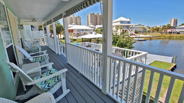 A covered porch with white railings, wicker furniture, and a view of a water body and buildings in the background. - Sunset Daze (Gulf Shores)