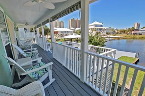 A covered porch with white railings, wicker furniture, and a view of a water body and buildings in the background. - Sunset Daze (Gulf Shores)