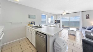 A modern kitchen with a dishwasher, a dining area with a table and chairs, a sofa, and a view of the sea. - Tradewinds 901 (Orange Beach)
