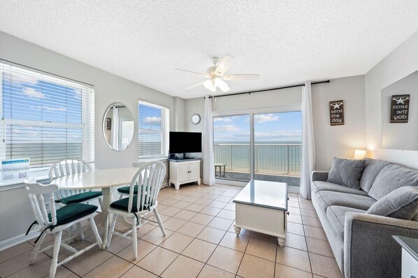 A spacious living area with a balcony, a dining table with chairs, a sofa, a television, and a ceiling fan.