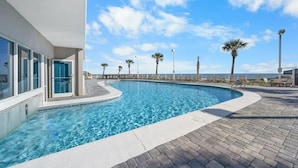 A swimming pool with clear blue water, surrounded by a paved deck and palm trees, with a building on one side. - Tradewinds 901 (Orange Beach)